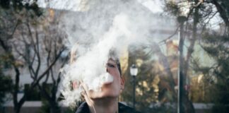 Photo of a teenager smoking, exhaling a large puff of smoke that almost completely covers his face.